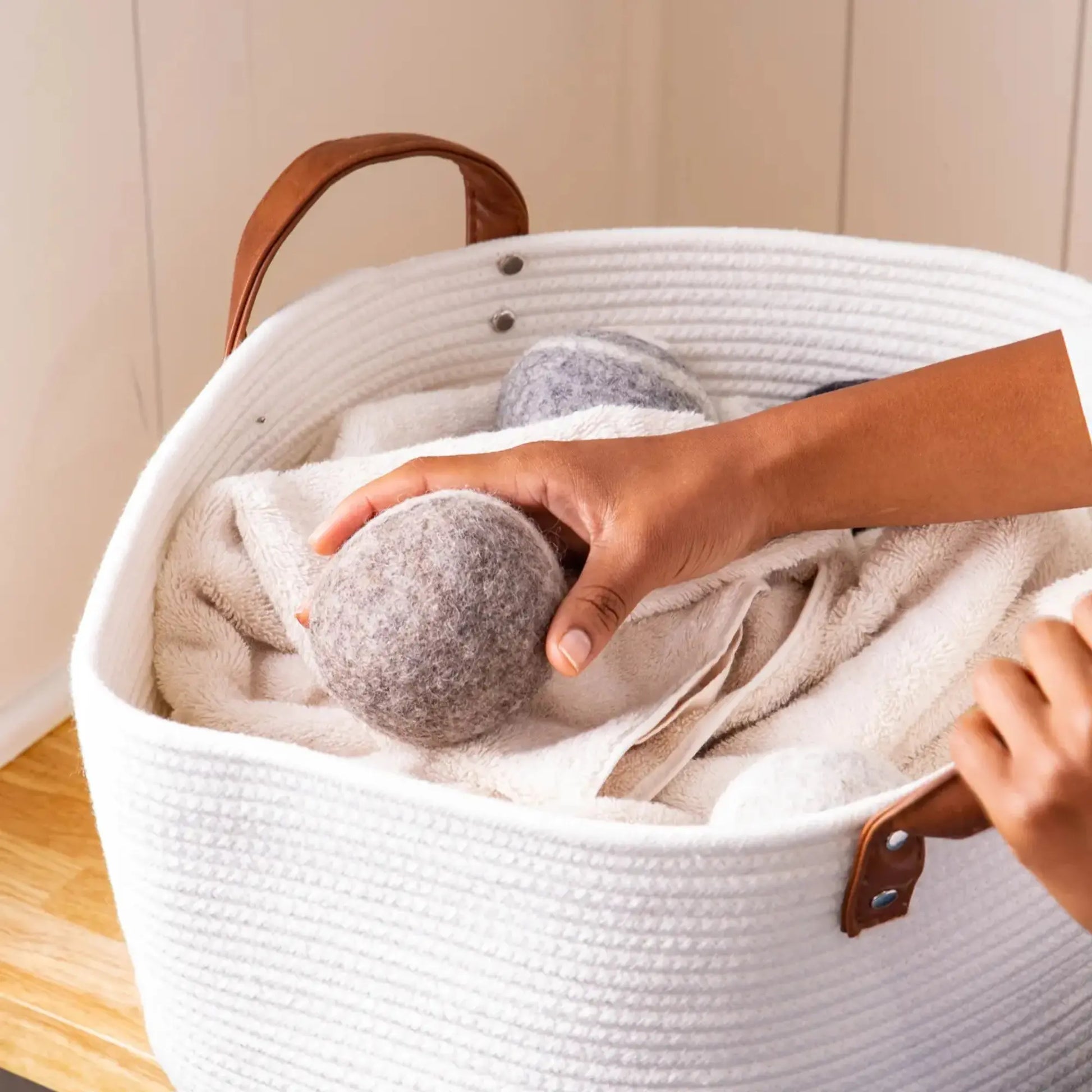 Person placing a gray dryer ball into a white laundry basket with brown handles.