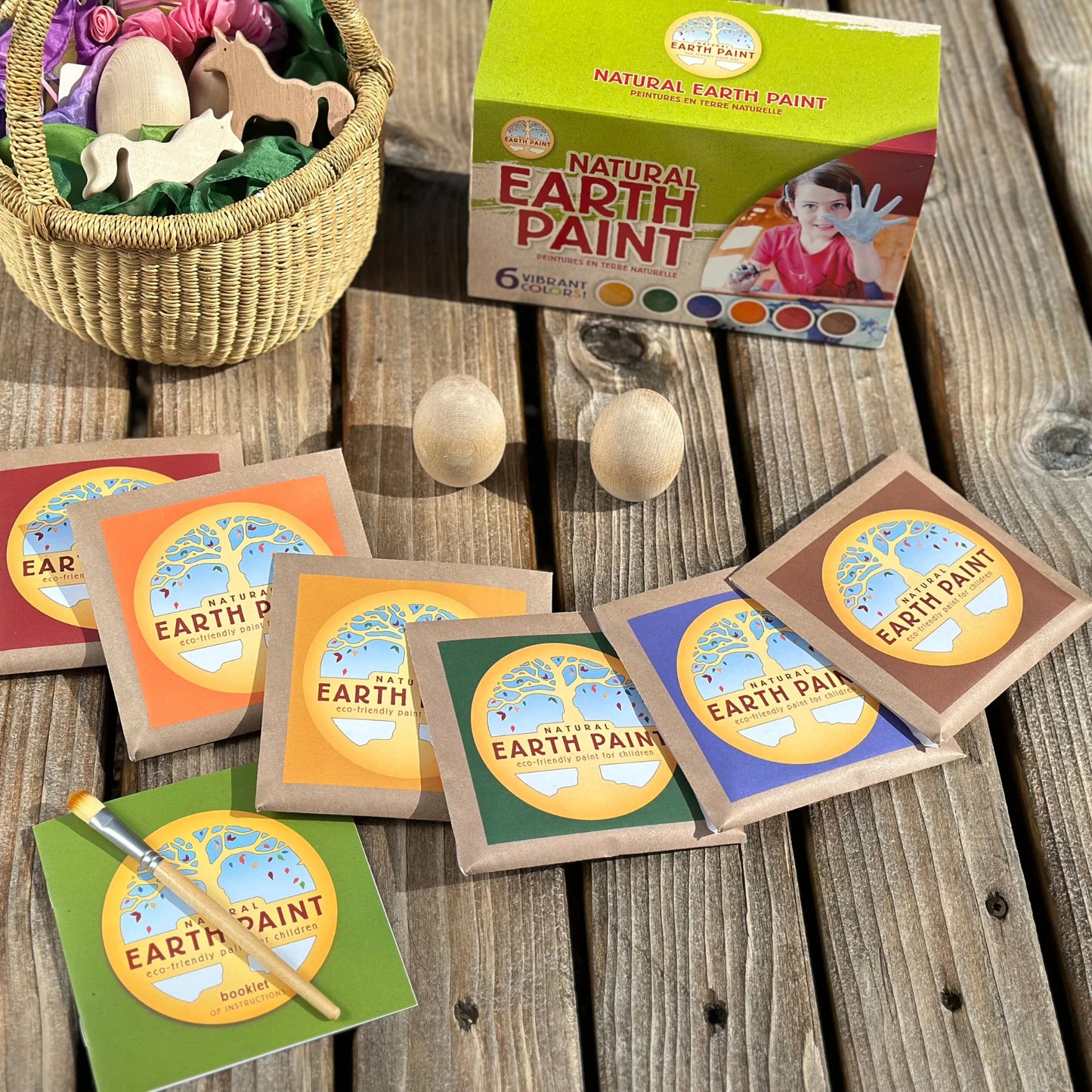 Earth paint packaging and samples on a wooden surface with a basket of wooden toys.