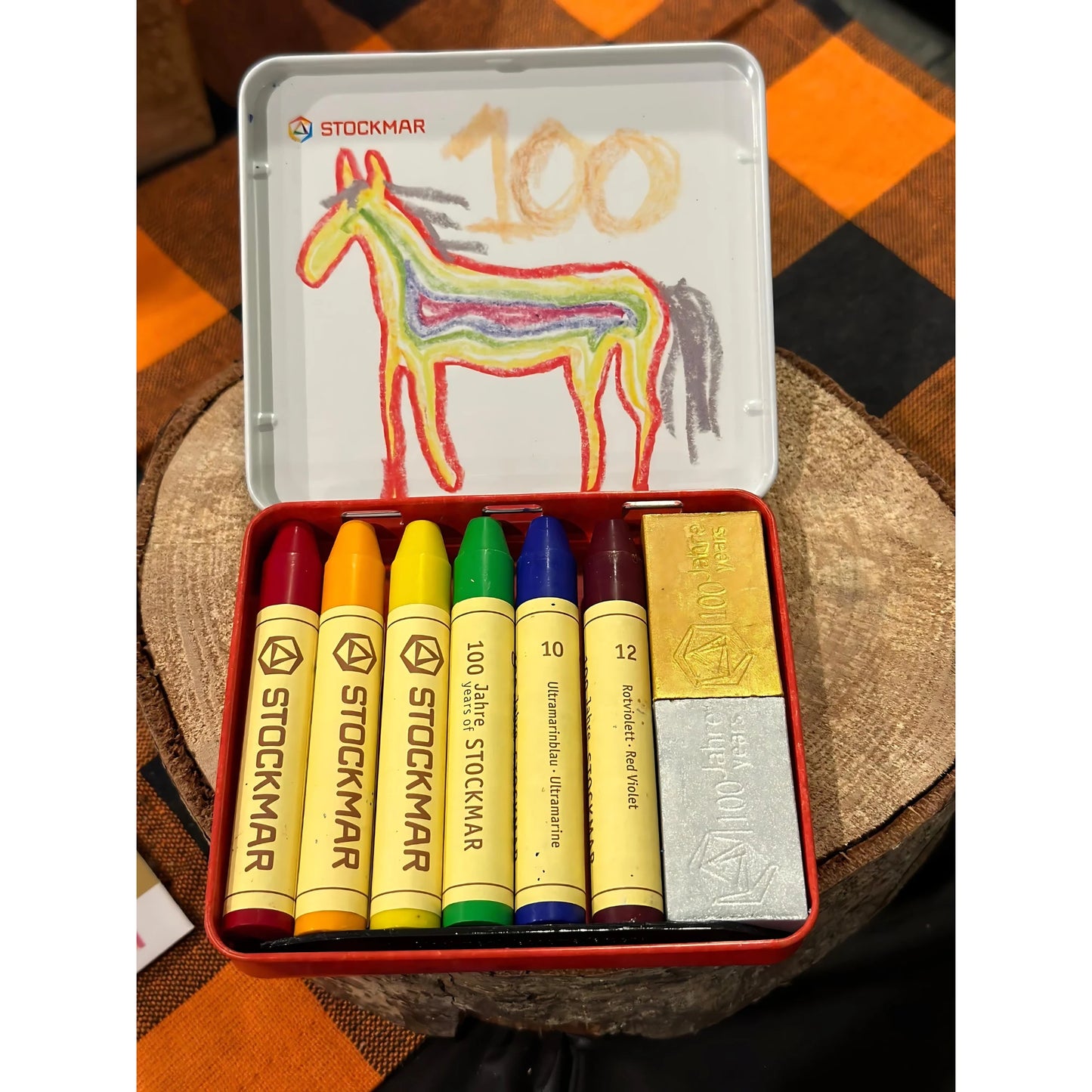 Set of Stockmar crayons in a tin with a child's drawing of a horse on a checkered fabric background.