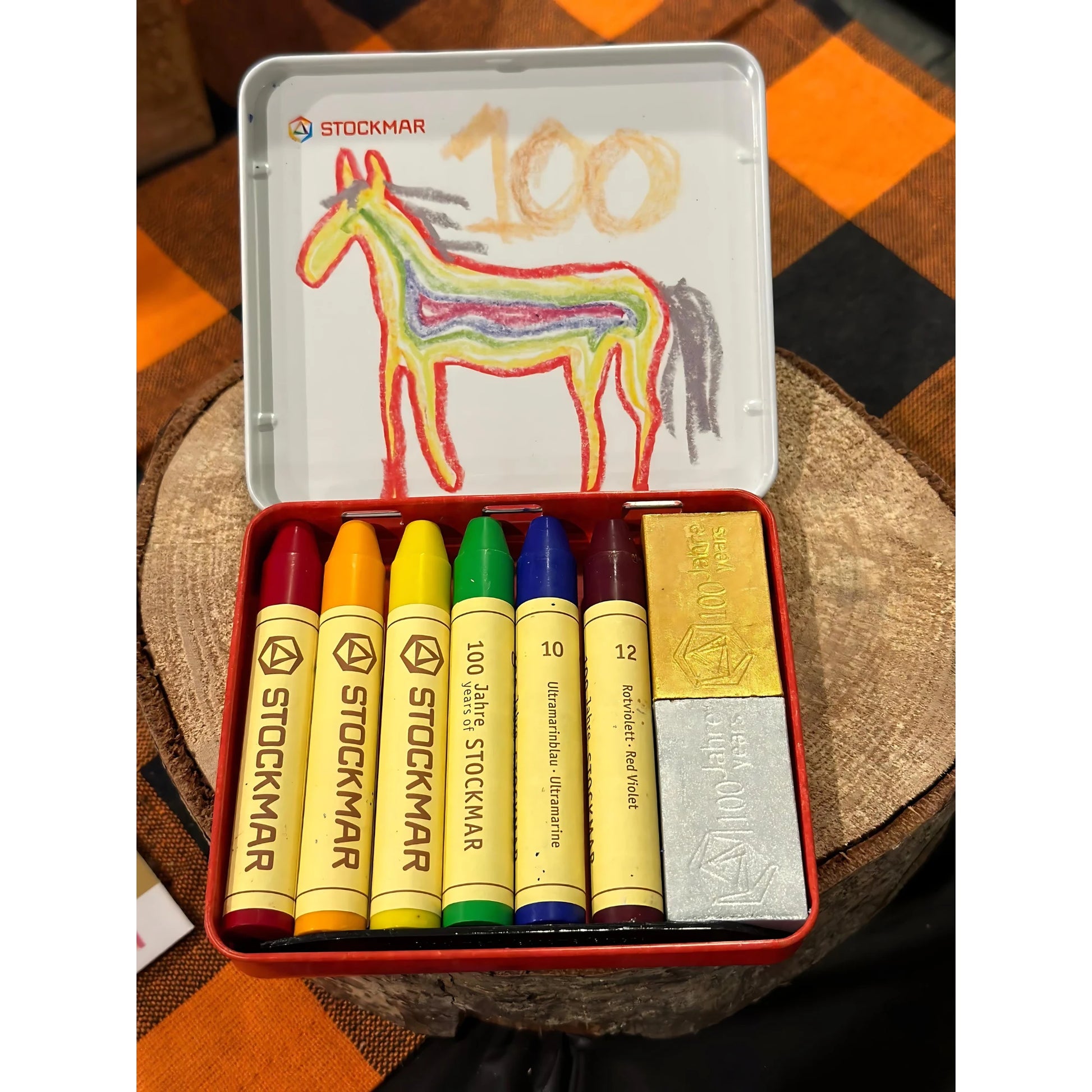 Set of Stockmar crayons in a tin with a child's drawing of a horse on a checkered fabric background.