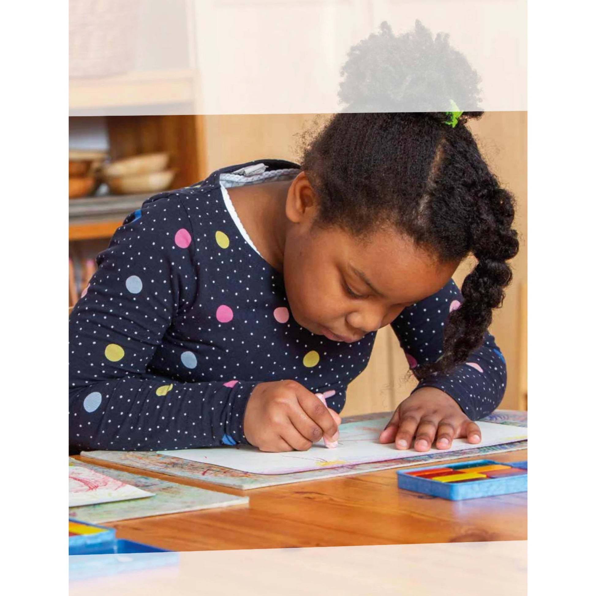 Child in a polka dot shirt coloring at a table with Stockmar crayons
